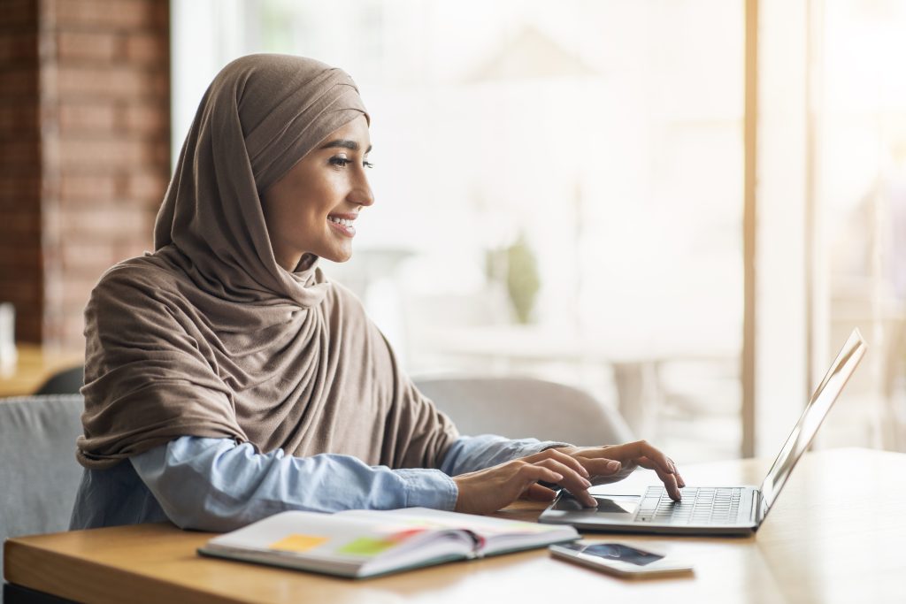 Cheerful girl in hijab sitting at cafe next to window, looking for job online, using laptop, side view, empty space